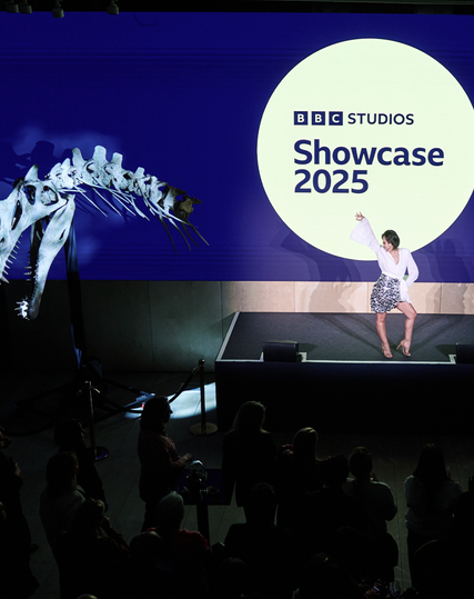 Photo of the stage at the BBC Showcase 2025. A reconstructed skeleton of the head and neck of the dinosaur Spinosaurus aegyptiacus is mounted in front of a large screen that displays the logo of the event.