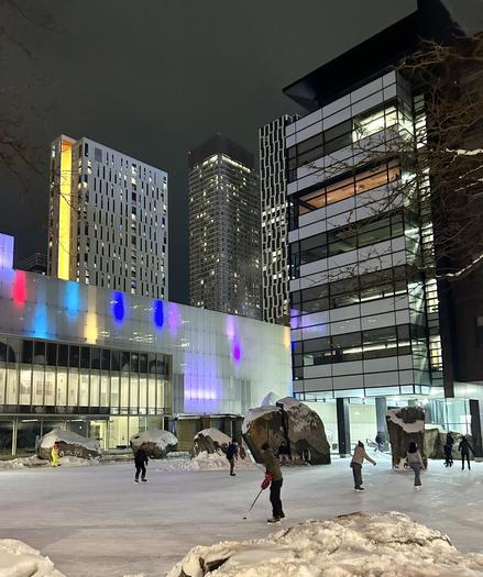 An illuminated ice skating rink is bustling with skaters at night, surrounded by modern buildings and snow-covered rocks. Colorful lights are reflected on the glass structures, enhancing the winter atmosphere.