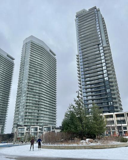 Two individuals ice skating on a snowy surface in an urban area, surrounded by tall residential buildings and a few trees. The sky is overcast and gray.