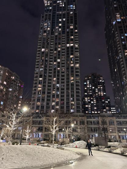 A person is ice skating on a path in a snowy area surrounded by tall, brightly lit buildings at night. Trees with lights are visible, along with snow piles and orange cones. The city skyline creates a vibrant urban atmosphere.