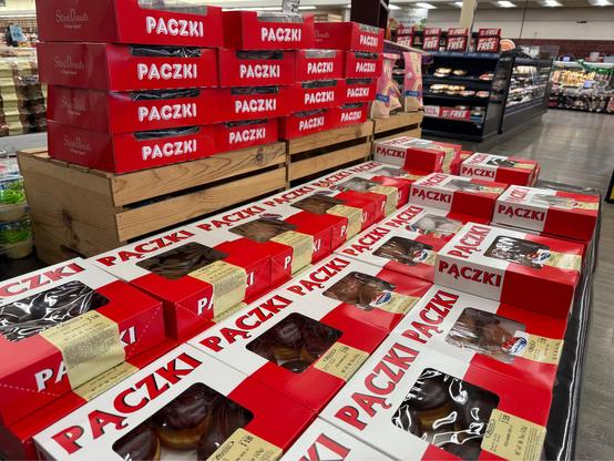 A display of Paczki, traditional Polish pastries, stacked in red and white boxes at a grocery store. The pastries are neatly arranged on wooden crates and tables, with some featuring clear plastic windows showing chocolate-covered varieties. The background includes store shelves with other baked goods and promotional signage.