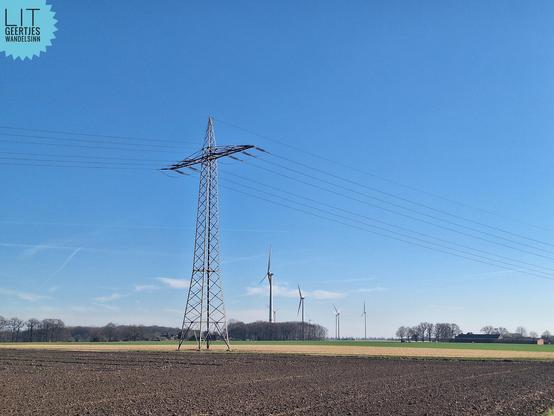 Landschaft mit braunen und gelben Feldern und Baumgruppen. Ein Strommast im Vordergrund, dahinter vier Windräder. Blauer Himmel, wenige Wölkchen. Sehr sonnig.