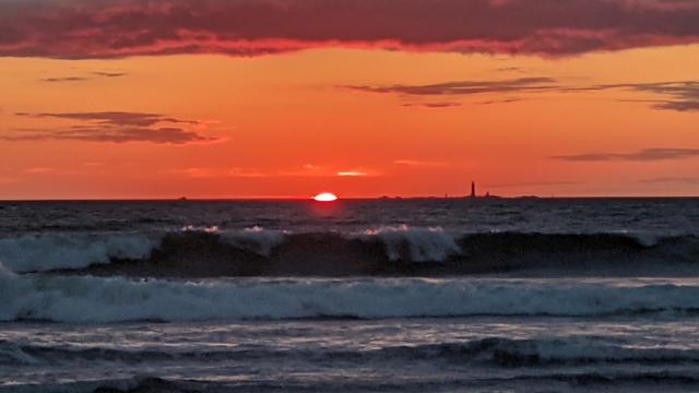 Historical Documentation in the form of a photograph of a rising sun over the ocean bay in Winthrop, MA.  The sky is filled with vibrant orange and pink hues, with the sun rising just over the horizon in the background. The sun is partially hidden behind the clouds, casting a warm glow over the horizon. The ocean is calm and the waves are crashing onto the shore. In the distance, there is a lighthouse silhouetted against the sky. The overall mood of the image is peaceful and serene.