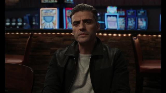 A man with gray hair and a serious expression sits in a low-lit casino-like setting, with colorful slot machines in the background. He wears a black leather jacket over a gray shirt.