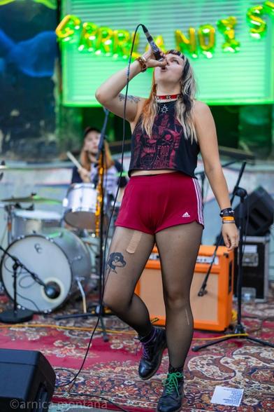 A performer sings passionately into a microphone on stage, wearing a black tank top and red shorts. She has torn fishnet stockings and black boots with green laces. In the background, a drummer is partially visible, and colorful lights illuminate the setting. 