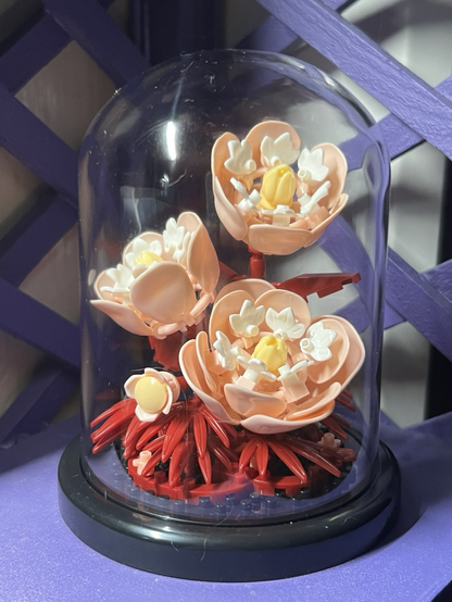 The finished flego set on my window sill. It consists of three big pale pink flowers with white and yellow accents sitting on a bed of dark red foliage, on a round black base with a protective clear display dome.