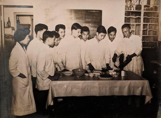 A sepia toned black and white image.  A group of Chinese people are standing around a long table.  A smiling culinary instructor appears to be pouring something out of a skillet into a bowl at one end of the table while a group of eight students watch attentively.  The author of this post's dad stands at the other end of the table facing the instructor.  A woman in a long white lab coat stands behind him with her hands in her pockets.