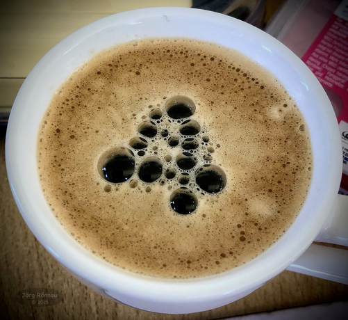 Das Bild zeigt eine Tasse Kaffee von oben. Die Oberfläche des Kaffees ist mit vielen kleinen und einigen größeren Blasen bedeckt. Die größeren Blasen bilden in der Mitte der Tasse die Zahl 4. Der Kaffee selbst sieht dunkelbraun und cremig aus. Die Tasse ist weiß.