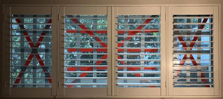 Four indoor plantation shutters covering windows, whose glass has been reinforced with gaff tape. The tape is applied in diagonal crosses, overlaid with a grid.