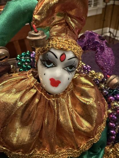 A close-up photograph of a decorative jester doll features a porcelain-like face with dramatic makeup, including arched black eyebrows, long eyelashes, dark eyeshadow, and red lips. A red jewel is painted on its forehead. The doll wears a metallic gold ruffled collar and a multicolored jester hat with green, gold, and purple fabric, adorned with small bells. In the background, Mardi Gras-style beaded necklaces in purple, green, and gold are visible, enhancing the festive theme. The setting appears to be indoors with warm lighting.