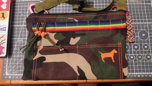 A small homemade bag sits on a cutting mat on a desk. The bag is camo cloth but is embellished with rainbow webbing loops, neon orange stitching and a bright orange concretedog logo. 