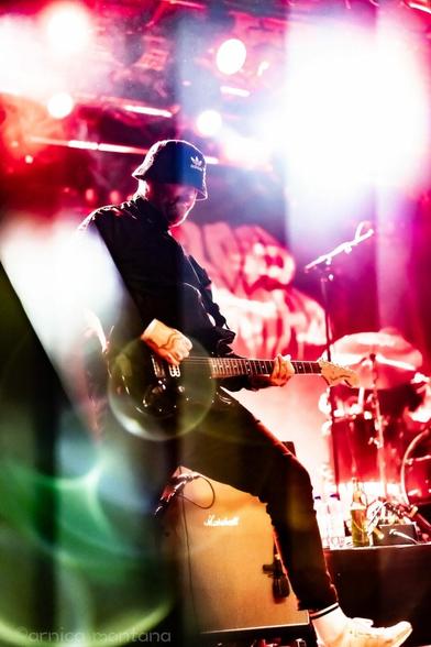 A guitarist performing on stage, wearing a bucket hat and a black outfit, with vibrant stage lights in red and white hues. A drum kit is partially visible in the background, and there is a Marshall amplifier nearby.