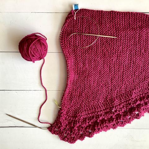 Flat lay view of a ball of dark pink yarn, and a knitting project featuring garter stitch and lace on a circular needle. It's shown on a white plank background.
