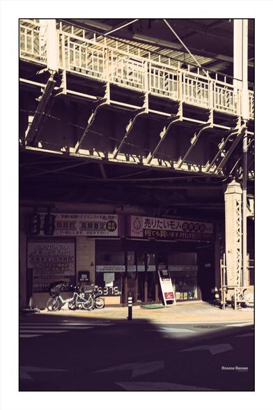 a photograph of under some transit infrastructure in Asakusa.  A big steel column holds up a train overpass, a small shop and some bicycles sit underneath.  The image is toned to look like film, highlights are a soft peach and shadows are warm.