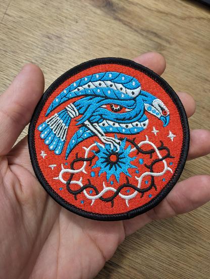 Patch brodé en rouge noir bleu représentant un oiseau tenu dans une main.