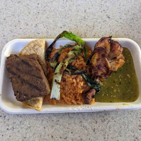 Grilled skirt steak on top of a small folded tortilla, next to a mound of Spanish rice with a grilled green onion on top, next to chipotle chicken, next to salsa verde.