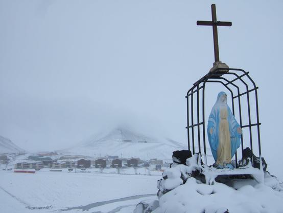In the foreground, a statue of the Virgin Mary is protected by a metal frame forming a curved arch.  A large crucifix is fastened to the top of the frame.  Some of the dormitory buildings of McMurdo Station are visible in the middle distance.  Observation Hill is partially visible behind "Mac Town".  The entire scene is covered in a light dusting of snow.