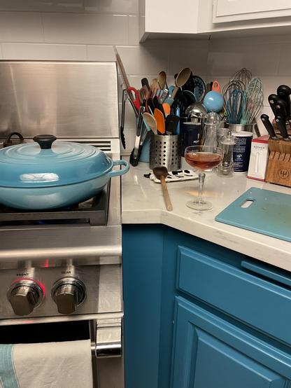 a corner of a kitchen.  A blue Le Creuset is on the stove, cooking jambalaya, and a coupé glass with a Sazerac is on the counter. A cutting board is visible, and there are several containers with cooking implements—spatulas, ladles, wooden spoons, tongs, as well as several types of cooking salt.