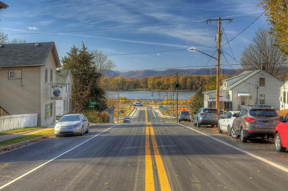 A quiet street stretches down toward a scenic river, flanked by parked cars and residential houses.