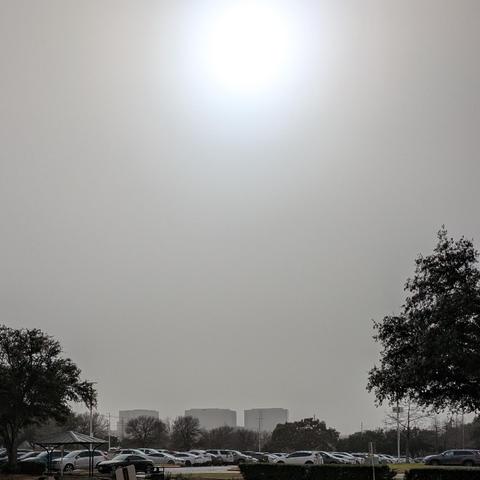 A photo showing three midrise office buildings in the distance behind a car-filled parking lot under the sun. There is a heavy haze across the whole scene.