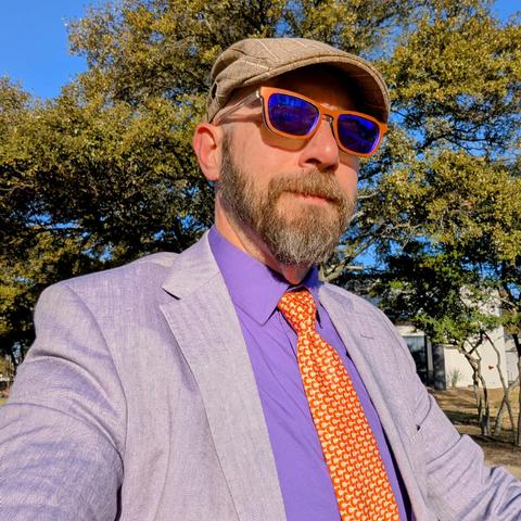 A photo of me, a 47 year old white man with a shaved head, a short cropped brown & grey beard, 

photographed from the chest up

In front of some live oak trees in an office park,

wearing

rectangular orange-yellow acetate glasses, a necktie with an orange crescent pattern in a trinity knot on a deep purple dress shirt under a lavender sport coat with a tan flat cap.