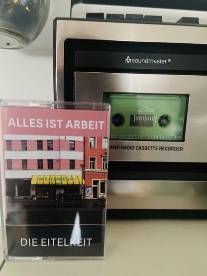 Bild einer Kassettenhülle mit dem Titel "ALLES IST ARBEIT" von "DIE EITELKEIT", neben einem Soundmaster-Kassettenrekorder. Der Hintergrund zeigt ein Gebäude mit einem Geschäft mit der Bezeichnung "BLUMEN".