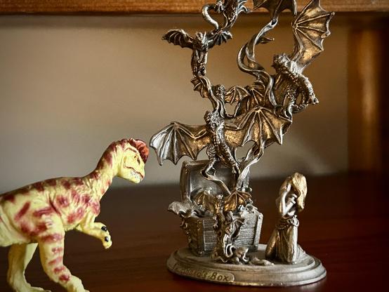 A green Dilophosaurus with brown spots watches in awe as a hoard of bat-like demons fly out of a opened chest. A pewter Pandora looks on in horror as the hell spawn take flight. Text reading "Pandora's Box" is visible on the bottom of the pewter statue. All are standing on a dark brown wooden surface.