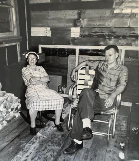 A grandmother sitting in a dress and apron, arms crossed knees wide. A man sitting legs crossed 