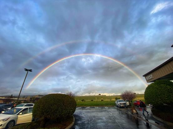 A wide view of sky with double rainbows stretching end to end . Foreground is parking area. 