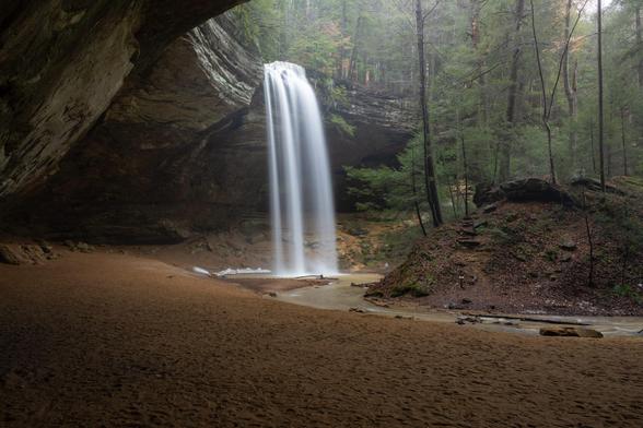 A waterfall at Ash Cave, a large recessed cave. The waterfall is high flow as it drops off the nearly 100' drop with a soft, misty shrouded trees off to the right and a sandy foreground.