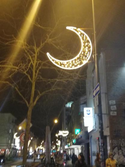 Blick auf eine abendliche erleuchtete Einkaufsstraße mit in regelmäßigen Abständen aufgehängten goldenen Leuchtdekorationen, die vorderste in Halbmondform