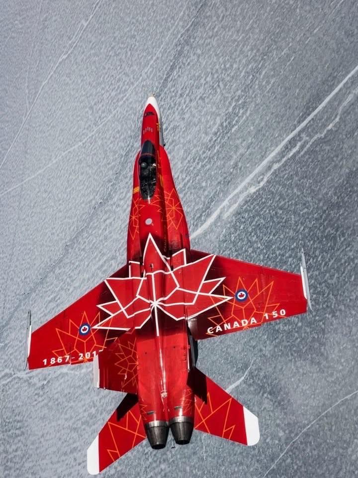 Une vue aérienne d'un avion de chasse rouge CF-18 Hornet, orné d'un motif de feuille d'érable canadien et du texte "CANADA 150" et "1867-2017". Le jet est positionné au-dessus d'une surface enneigée, mettant en valeur ses couleurs vives sur le fond blanc. Une vue aérienne d'un avion de chasse rouge CF-18 Hornet, orné d'un motif de feuille d'érable canadien et du texte "CANADA 150" et "1867-2017". Le jet est positionné au-dessus d'une surface enneigée, mettant en valeur ses couleurs vives sur le fond blanc.