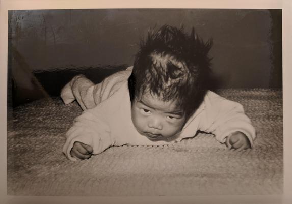 A black and white photo of a baby sprawled on a bed, fists clenched, head tilted up, starting down the camera.