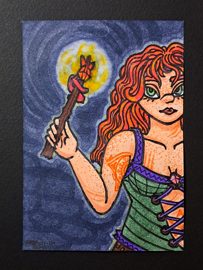 Un dessin représentant Adeline, une femme rousse aux yeux bleus portant un corset vert bordé de violet avec des lacets de la même couleur, ainsi qu'un pantalon de cuir ; elle brandit la Baguette des Flammes, une baguette de bois sombre d'environ 40cm orné d'un crystal orangé à 3 pointes, avec une aura enflammée autour.