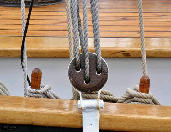 Das maritime Bild zeigt einen Ausschnitt vom Deck eines historischen Segelschiffes. Im Mittelpunkt befindet sich ein hölzerner Taljenblock in der Takelage, durch den mehrere Taue laufen. Links und rechts davon sind weitere Taue an hölzernen Belegnägeln befestigt. Das Deck des Schiffes besteht aus hellem Holz. 