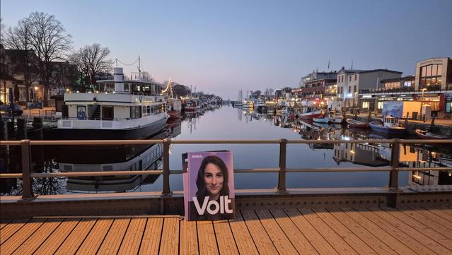 Warnemünde Alter Strom mit sich im Wasser spiegelnden Gebäuden und Volt Plakat im Vordergrund