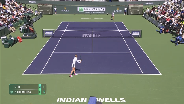 Screenshot of Russian tennis player Polina Kudermetova serving in the near court on the ad side, to American Claire Liu. An on-screen scorecard in the lower-left shows the score as 0-3 15-0 in the 1st set. She wears a white quarter-zip long-sleeved athletic top with EA7 logos, an off-white tennis skirt with a dark-blue side stripe, white & dark-pink sneakers, small earrings, black bracelets on her left writst and what looks like a white sweatband & bracelet on her right wrist, and has her blonde hair in a bun at the back of her head. Her tennis racquet is a dark-green Wilson with a white grip. Her opponent, Claire Liu, wears a lightweight ASICS white quarter-zip long-sleeved athletic top with black collar and what looks like integrated black sweatbands, a pleated grayed-purple tennis skirt, pink sneakers, pearl earrings, a white sweatband on her left wrist, a white visor, and has her black hair in a bun behind her head. Liu has an orange Head racquet with white grip. The court is blue and the out-of-bounds area is green. A rectangular blue sign with the Holologic logo in white is at each end of the net. The baseline wall is dark green and shows the tournament name and sponsor logos. On-court baseline logo reads "Indian Wells" in white block letters. Ball kids wear light-gray tournament uniforms. The chair umpire is at left center-court on a shaded, elevated dark-green seat, and wears a black uniform. The on-court bleachers on either side are mostly full with spectators.