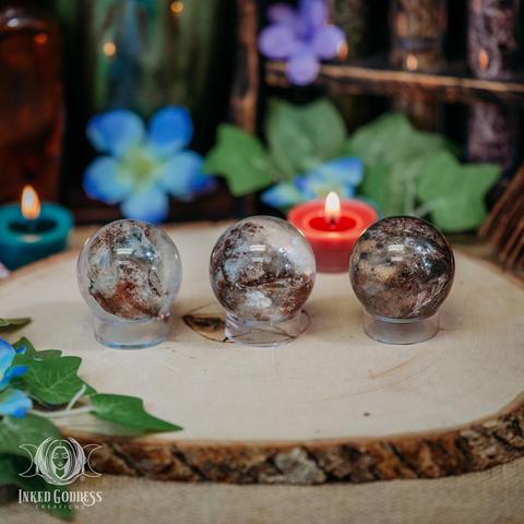 Three specimens of the "Garden Quartz / Lodolite Gemstone Sphere" product are arranged on a raw wood slice. The spheres are about 1.5 inches in diameter and have sections of transparent quartz with opaque areas of brown lodolite.. Presented by Inked Goddess Creations.