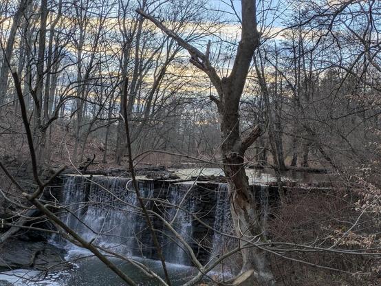 Photo of a waterfall along a creek in the woods. The trees are bare. The sky is blue and streaked with clouds.