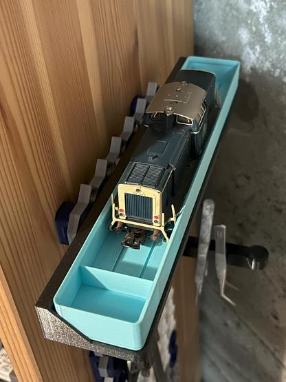 Picture of a Marklin model train locomotive in a blue plastic Gridfinity cradle, hanging from a Multiboard pegboard, which is attached to a bookcase.