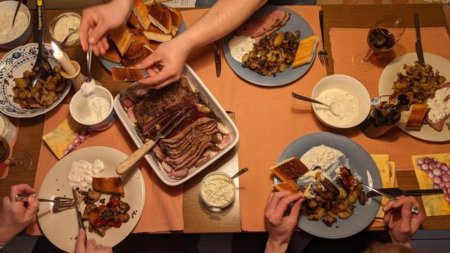 Bildbeschreibung von KI:
Das Foto zeigt eine gemütliche Essensszene aus der Vogelperspektive. Mehrere Personen sitzen an einem gedeckten Tisch und genießen eine herzhafte Mahlzeit.  

In der Mitte des Tisches befindet sich eine große Auflaufform mit aufgeschnittenem, rosa gegartem Fleisch, das mit Kräutern und Knoblauch gewürzt wurde. Dazu gibt es geröstete Kartoffeln mit Gemüse, verschiedene Dips oder Saucen sowie knuspriges Brot. Jeder Gast hat einen eigenen Teller mit Portionen der Speisen, und man sieht mehrere Hände, die aktiv essen oder sich Essen nehmen.  

Die Atmosphäre ist warm und gesellig, mit einer brennenden Kerze als Dekoration. Getränke wie Rotwein und Bier ergänzen das Mahl. Die Tischdekoration besteht aus orangefarbenen Platzdeckchen und Servietten mit einem Traubenmotiv. Das Bild vermittelt das Gefühl eines gemeinsamen, genussvollen Abendessens mit Freunden oder Familie.