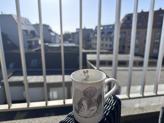 A white ceramic mug with a black-and-white sketch of a skeptic Mickey Mouse sits on a woven outdoor chair. In the background, a parking area with multiple garage doors is visible, along with modern residential buildings. White vertical bars of a balcony railing frame the scene, and sunlight casts a bright glow over the area.