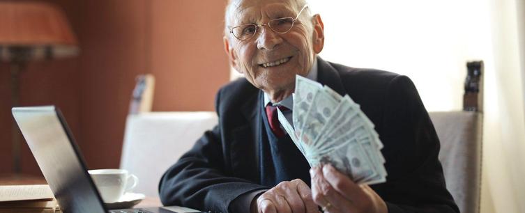 An elderly man smiling while holding a fan of cash, sitting at a desk