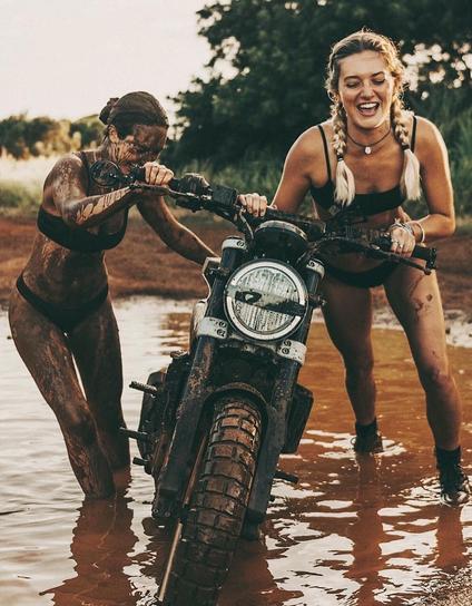 Two laughing white women in bikinis pushing a motorcycle through the mud. They're also pretty dirty with mud themselves.