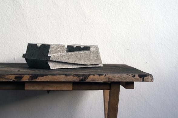 Side shot from an abstract geometric shaped ashtray. The ashtray is from concrete and is sanded down so specs of crushed shells and river sand appear on the surface resulting in a Terrazzo finish. The ashtray stands on a wooden table spattered with black paint.