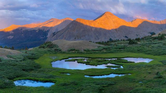 A high-altitude alpine scene with a series of reflective ponds surrounded by lush green vegetation in the foreground. Rolling hills and rugged mountain peaks rise in the background, bathed in warm, golden light from the setting or rising sun. Shadows stretch across the landscape, contrasting with the vibrant tones of the illuminated peaks. The sky is a mix of soft blues and grays, with clouds adding depth to the composition.
