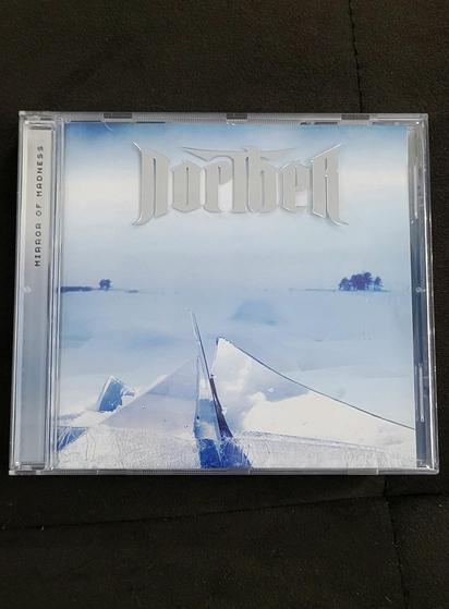 A CD album on dark brown background. The whole cover is in blueish white tone. A horizon can be made out in the middle, with some darker blue trees near the left and right border. In the foreground there are several big shards of glass covered in snow and loosely arranged to form a pyramid shape. The name of the band Norther is embossed in silver characters on the plastic cover itself, while the album name "Mirror of Madness" is only on the top of the side hinge in old school computer game font.