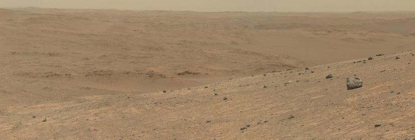 An ochre toned picture of a Martian landscape. A standalone grey colored rock stands out on the slope of the rim of the crater on the right hand side. 