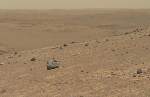 An ochre toned picture of a Martian landscape. A standalone grey colored rock stands out on the slope of the rim of the crater in the foreground. 