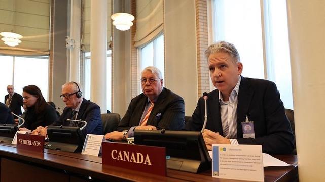Ambassador Hugh Adsett of Canada speaks at the 108th session of the OPCW’s Executive Council. He sits behind the Canada nameplate in the Ieper Room at OPCW HQ, and his Dutch colleague listens beside him. 